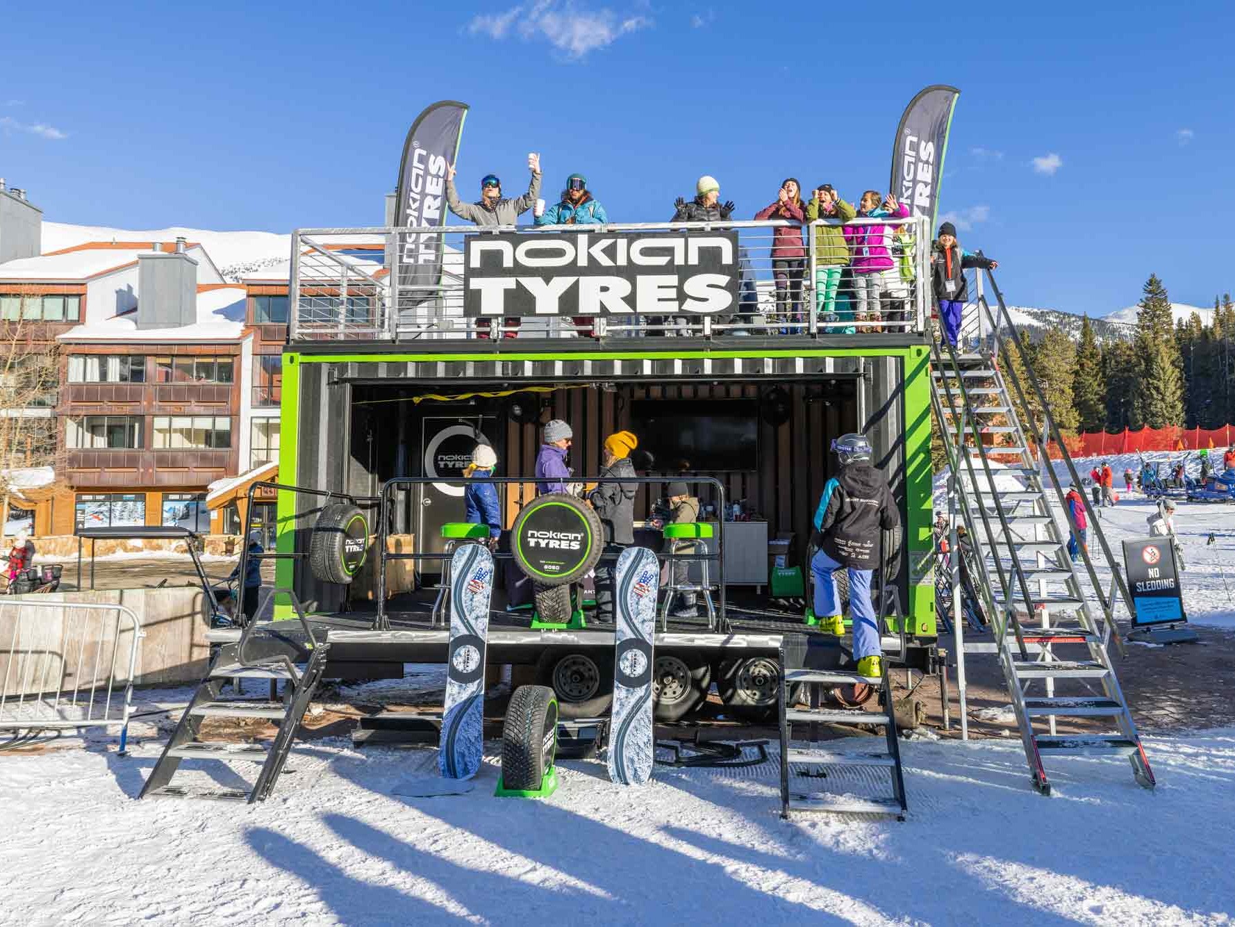 A Nokian Tyres promotional booth with banners is set up in a snowy ski resort area, highlighting the durability of their tyres even in winter conditions. People stand on and around the booth, with ski equipment and snow visible in the background, while a Roadie Pro display showcases Nokian Tyres’ commitment to safety on icy roads.