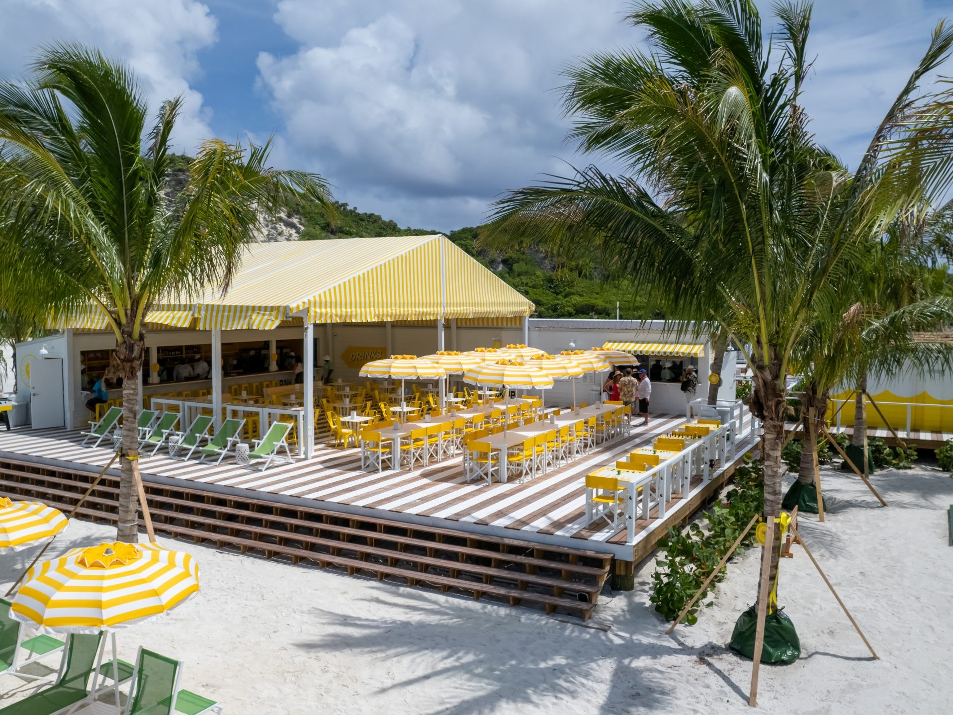 A beachside café with yellow-and-white striped awnings, tables, and umbrellas, surrounded by palm trees on white sand under a partly cloudy sky. Green lounge chairs are arranged near the seating area, giving this inviting spot the relaxed charm often found in light commercial building construction types along the coast.