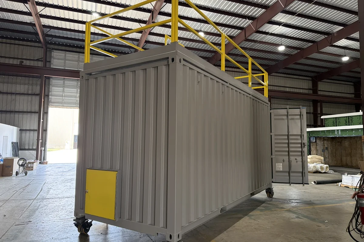 A large gray metal container with yellow railings on top and a yellow door sits inside a spacious industrial warehouse with high ceilings and exposed beams.