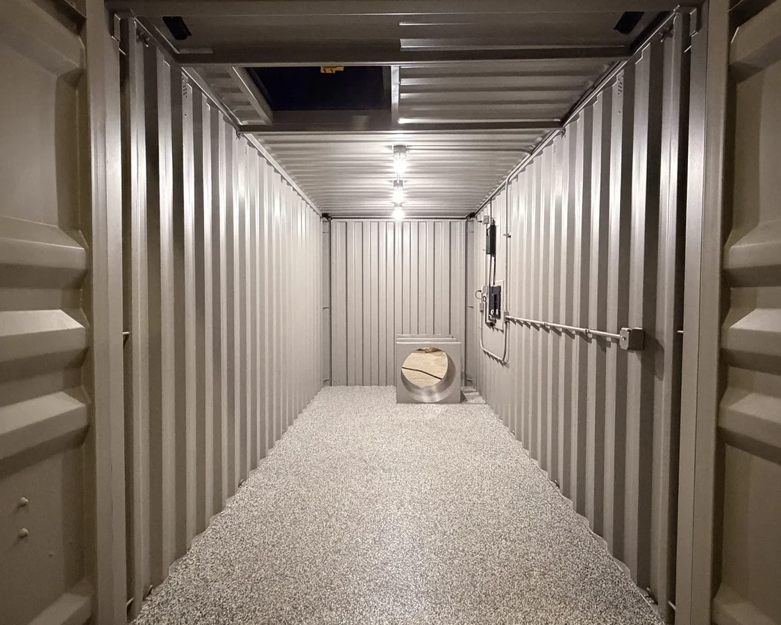 Interior of an empty, metal shipping container with lights on the ceiling, a small square opening near the top, and a circular metal structure partially covered with beige fabric at the far end. Pebbled flooring is visible.