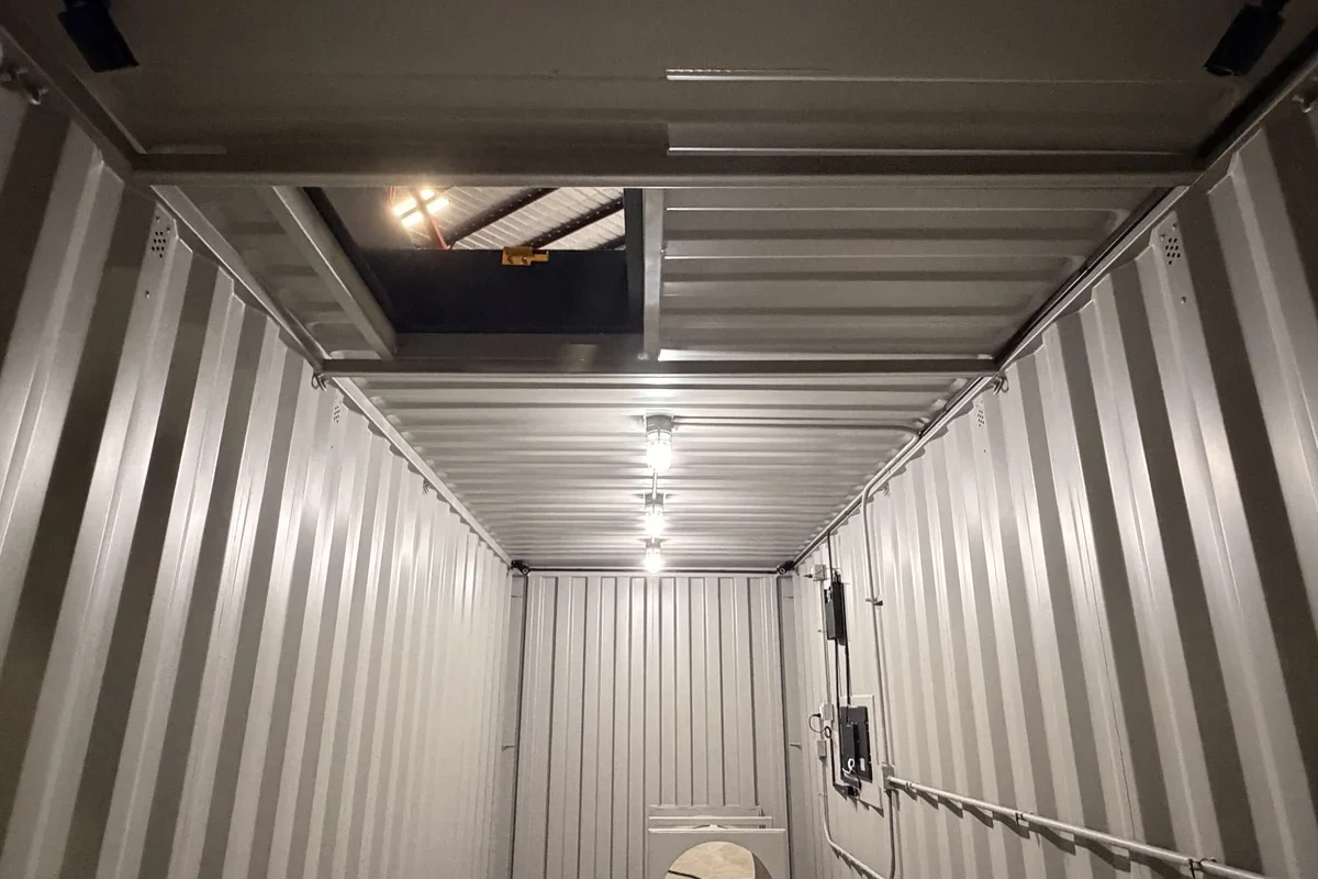 Interior of a metal shipping container with corrugated walls, a ceiling hatch open to an overhead space, ceiling lights, visible wiring, and a small electrical panel on the right wall. A circular object sits near the back wall.